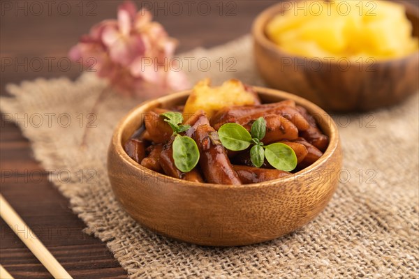 Tteokbokki or Topokki, fried rice cake stick, popular Korean street food with spicy jjajang sauce and pineapple on brown wooden background and linen textile. Side view, selective focus