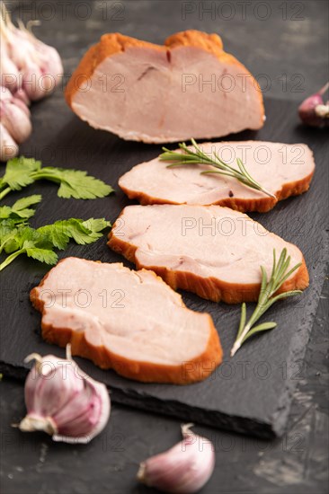 Smoked pork ham on cutting slate board with pepper, garlic and herbs on black concrete background. Side view, close up, selective focus