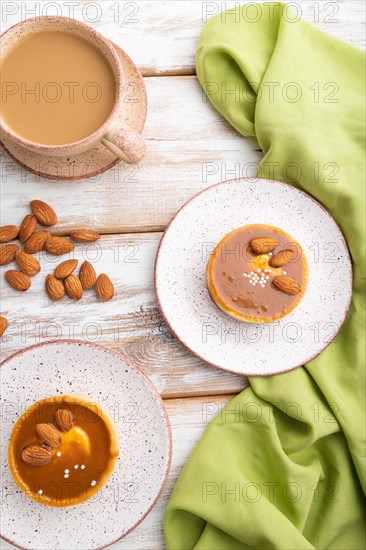 Sweet tartlets with almonds and caramel cream with cup of coffee on a white wooden background and green textile. top view, flat lay, close up