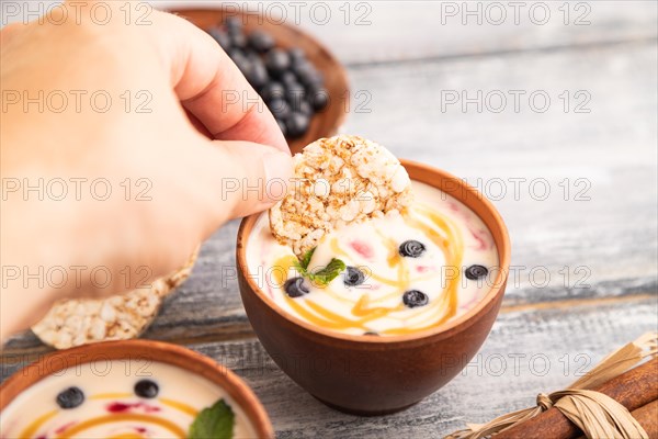 Yoghurt with bilberry and caramel in clay bowl on gray wooden, hand dipping cookies in yogurt, side view, close up, selective focus