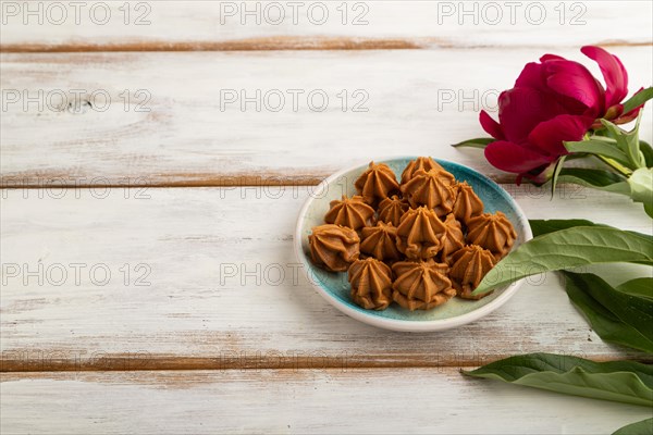 Homemade soft caramel fudge candies on blue plate on white wooden background, peony flower decoration. side view, copy space