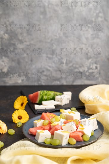 Vegetarian salad with watermelon, feta cheese, and grapes on blue ceramic plate on black concrete background and yellow linen textile. Side view, copy space, selective focus