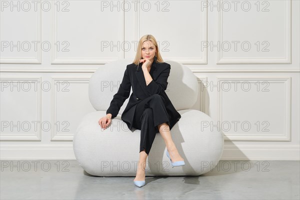 Positive businesswoman in casual suit sits on large bean bag propping chin and looking with interest