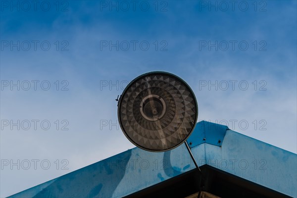Large street light with a metal shade mounted to the roof of a metal building with blue sky in the background