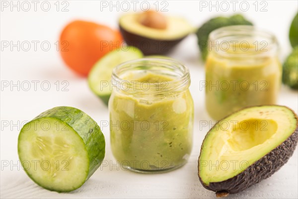Baby puree with vegetable mix, broccoli, tomatoes, cucumber, avocado infant formula in glass jar on white wooden background. Side view, close up, selective focus, artificial feeding concept
