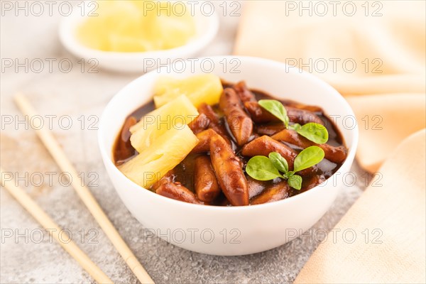 Tteokbokki or Topokki, fried rice cake stick, popular Korean street food with spicy jjajang sauce and pineapple on gray concrete background and orange textile. Side view, close up, selective focus