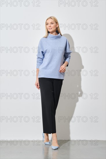 A modern fashion model poses in a relaxed stance, wearing a stylish blue sweater paired with classic black trousers, complemented by light blue heels