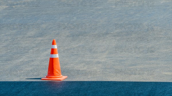 Orange traffic cone sitting in a pave parking lot on a sunny day with a shadow of a building in bottom of frame