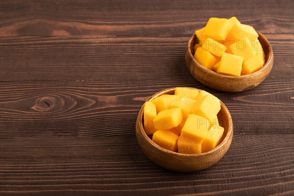 Dried and candied mango cubes in wooden bowls on brown wooden textured background. Side view, copy space, vegan, vegetarian food concept