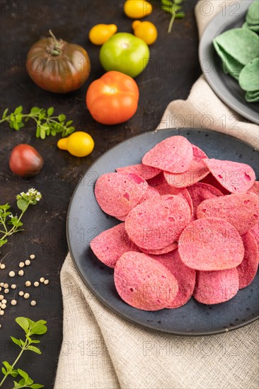 Green and red potato chips with herbs and tomatoes on black concrete background and linen textile. Side view, close up, selective focus