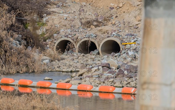 Three water drainage culverts in side of hill that empty into shallow river. Bridge support column blurred in foreground