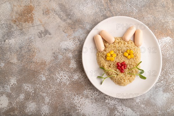 Funny mixed quinoa porridge, sweet corn, pomegranate seeds and small sausages in form of cat face on brown concrete background. Top view, flat lay, copy space. Food for children, healthy food concept