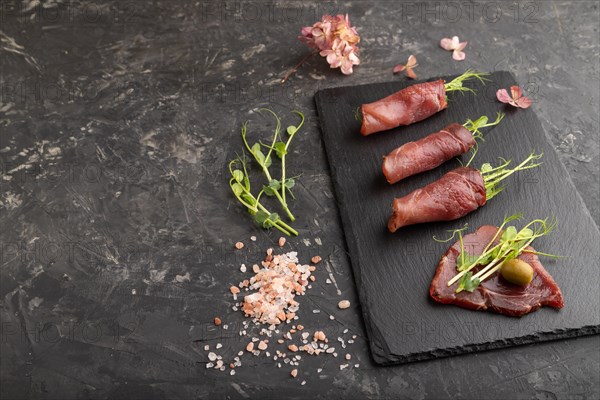 Slices of smoked salted meat with green pea microgreen on black concrete background. Side view, copy space