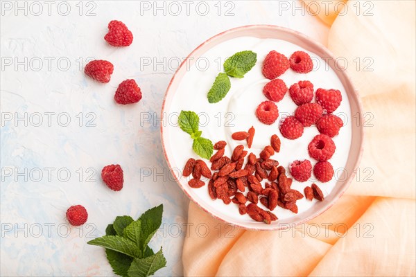 Yogurt with raspberry and goji berries in ceramic bowl on white concrete background and orange linen textile. top view, flat lay, close up