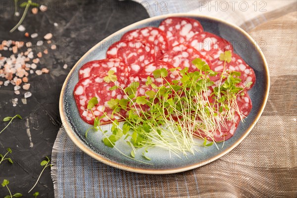 Slices of smoked cervelat salami sausage with spinach microgreen, salt and pepper on black concrete background and beige textile. Side view, close up, selective focus