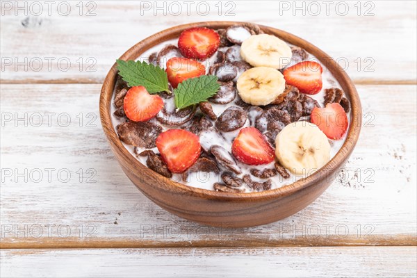 Chocolate cornflakes with milk and strawberry in wooden bowl on white wooden background. Side view, close up