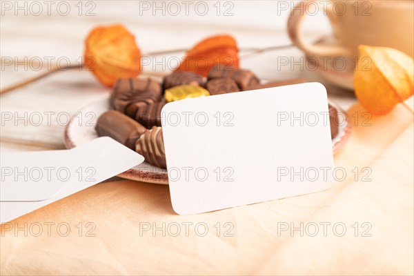 White business card with chocolate candies and physalis flowers on a white wooden background and orange textile. side view, close up, selective focus