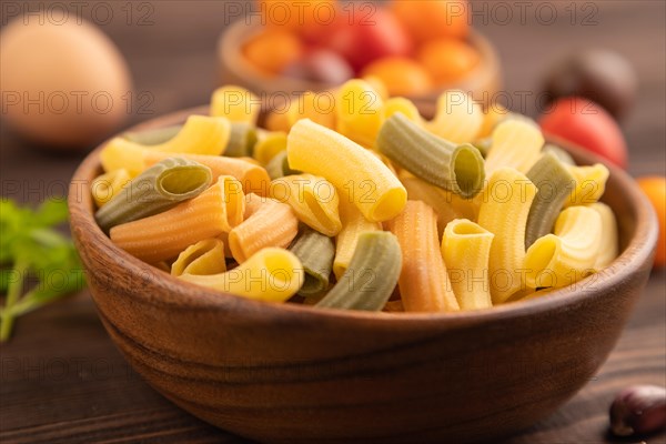 Rigatoni colored raw pasta with tomato, eggs, spices, herbs on brown wooden background. Side view, close up, selective focus