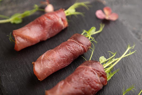 Slices of smoked salted meat with green pea microgreen on black concrete background. Side view, close up, selective focus