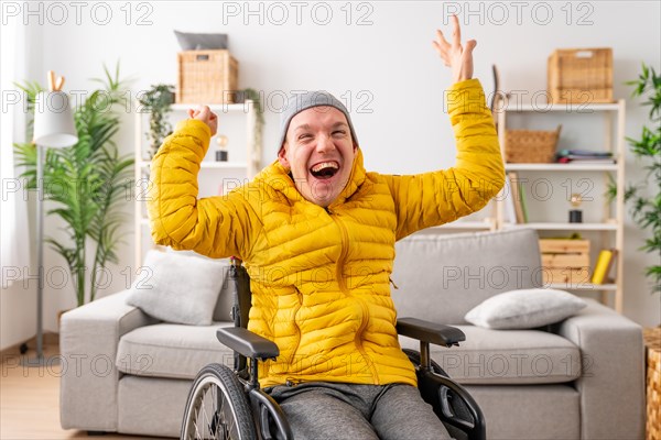 Portrait of a happy disabled man in wheelchair raising hands in joy celebrating in the living room at home