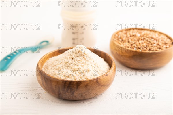 Powdered milk and buckwheat baby food mix, infant formula, pacifier, bottle, spoon on white wooden background. Side view, close up, selective focus, artificial feeding concept
