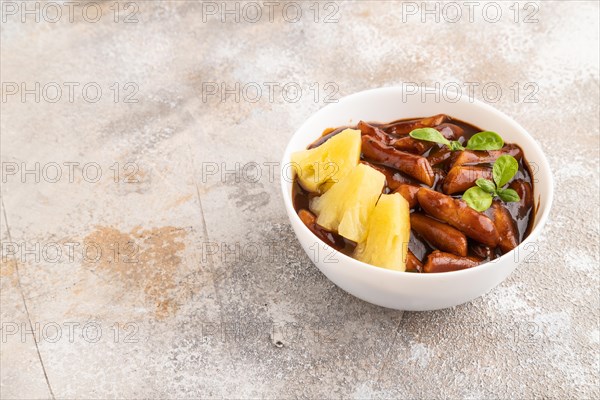 Tteokbokki or Topokki, fried rice cake stick, popular Korean street food with spicy jjajang sauce and pineapple on gray concrete background. Side view, copy space