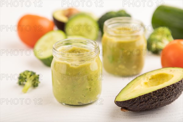 Baby puree with vegetable mix, broccoli, tomatoes, cucumber, avocado infant formula in glass jar on white wooden background. Side view, close up, selective focus, artificial feeding concept