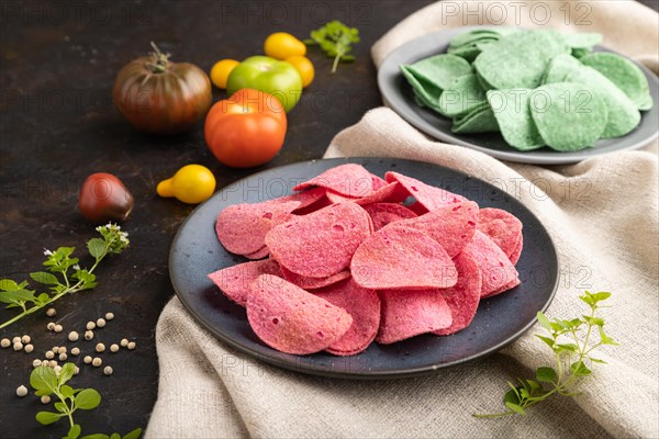 Green and red potato chips with herbs and tomatoes on black concrete background and linen textile. Side view, close up