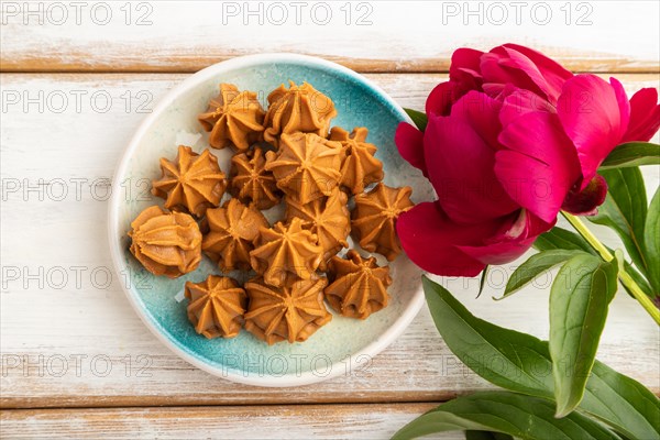 Homemade soft caramel fudge candies on blue plate on white wooden background, peony flower decoration. top view, flat lay, close up