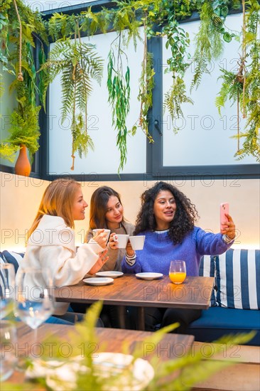 Vertical shot of three female friends taking selfie with mobile phone while drinking coffee in a cafeteria
