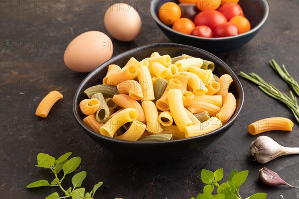 Rigatoni colored raw pasta with tomato, eggs, spices, herbs on black concrete background. Side view, close up