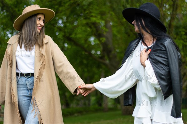 Front view of a stylish lesbian couple in hat holding hands and walking in the park while looking at each other