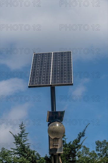 Small solar panel on top of light pole above treetops against blue cloudy sky