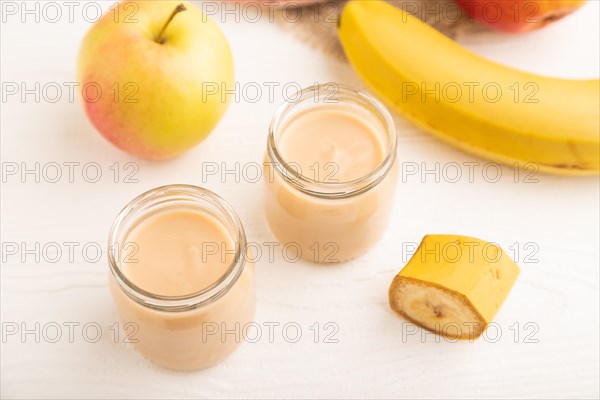 Baby puree with fruits mix, apple, banana infant formula in glass jar on white wooden background. Side view, close up, selective focus, artificial feeding concept