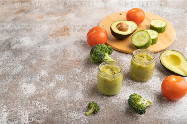 Baby puree with vegetable mix, broccoli, tomatoes, cucumber, avocado infant formula in glass jar on brown concrete background. Side view, copy space, artificial feeding concept