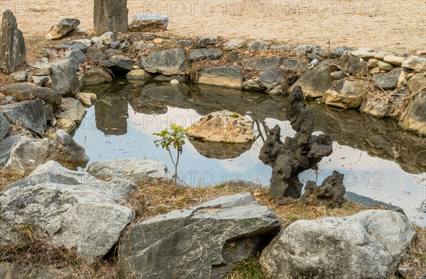 Piece of basalt shaped like tree next to small man made pond surrounded by granite boulders in South Korea