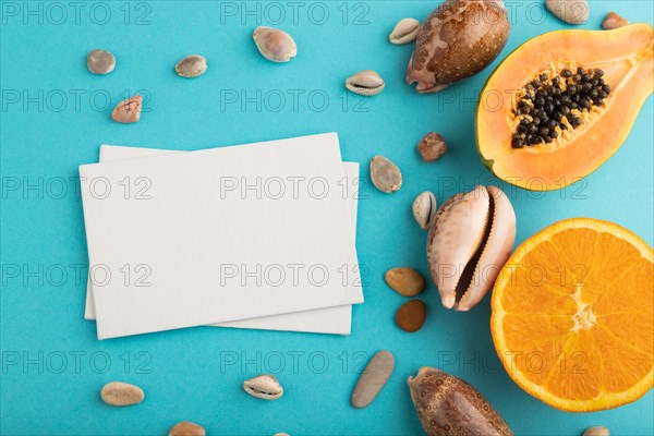 White paper business card with ripe cut papaya, orange, seashells on blue pastel pastel background. Top view, flat lay, copy space. Tropical, healthy food, vacation, holidays concept