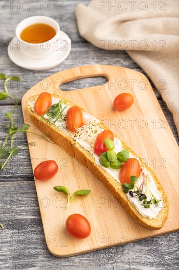 Long white bread sandwich with cream cheese, tomatoes and microgreen on gray wooden background and linen textile. side view, close up, selective focus