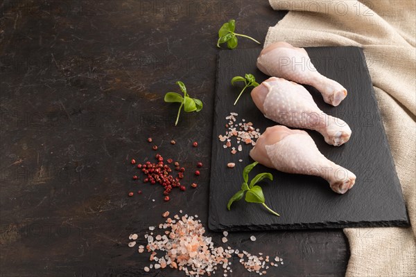 Raw chicken legs with herbs and spices on a black slate cutting board on a black concrete background and linen textile. Side view, copy space