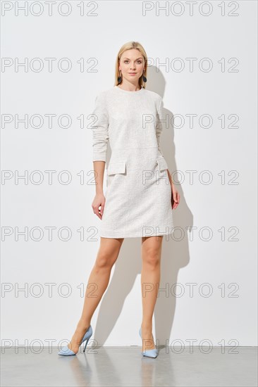 Cute woman in textured white mini dress with pockets and blue heels posing against a white wall