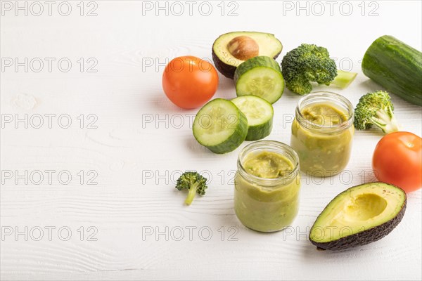 Baby puree with vegetable mix, broccoli, tomatoes, cucumber, avocado infant formula in glass jar on white wooden background. Side view, copy space, artificial feeding concept