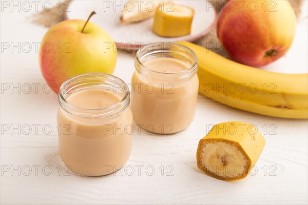 Baby puree with fruits mix, apple, banana infant formula in glass jar on white wooden background. Side view, close up, artificial feeding concept