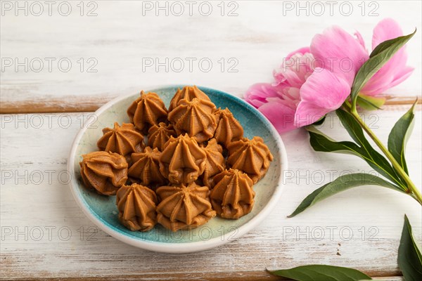 Homemade soft caramel fudge candies on blue plate on white wooden background, peony flower decoration. side view, close up