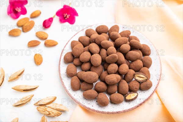 Almond in chocolate dragees on ceramic plate and a cup of coffee on white concrete background and orange linen textile. Side view, close up, selective focus