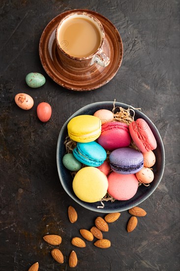 Multicolored macaroons and chocolate eggs in ceramic bowl, cup of coffee on black concrete background. top view, flat lay, close up, still life. Breakfast, morning, concept