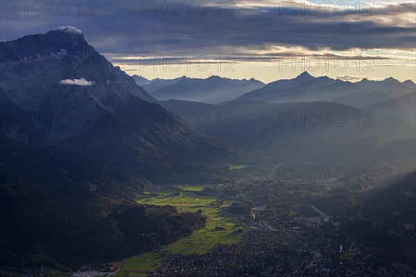 Distant view of town in front of mountains in the evening light, backlight, haze, Garmisch-Partenkirchen, left Zugspitze, Wetterstein Mountains, Bavarian Alps, Upper Bavaria, Bavaria, Germany, Europe