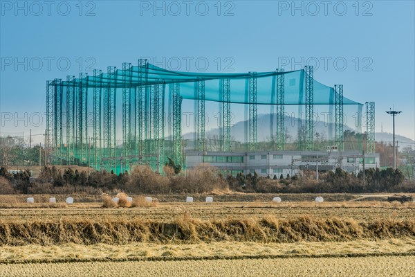 Golf driving range constructed of metal trussed columns and green netting in rural farming landscape with field of cut hay in foreground