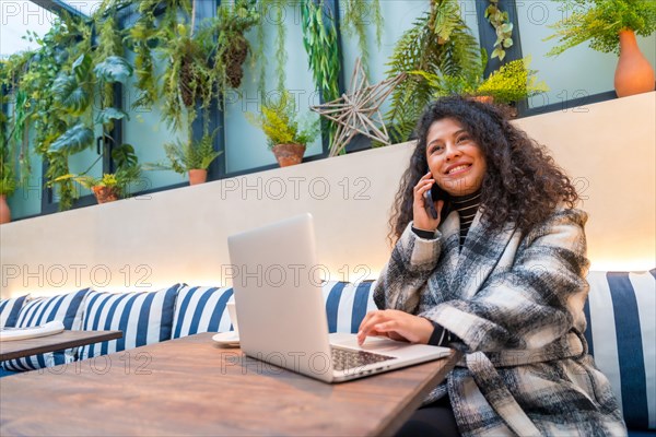 Smiling young woman with curly long hair using phone while working with laptop in a cafeteria