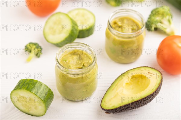 Baby puree with vegetable mix, broccoli, tomatoes, cucumber, avocado infant formula in glass jar on white wooden background. Side view, close up, selective focus, artificial feeding concept