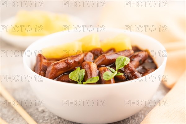 Tteokbokki or Topokki, fried rice cake stick, popular Korean street food with spicy jjajang sauce and pineapple on gray concrete background and orange textile. Side view, close up, selective focus